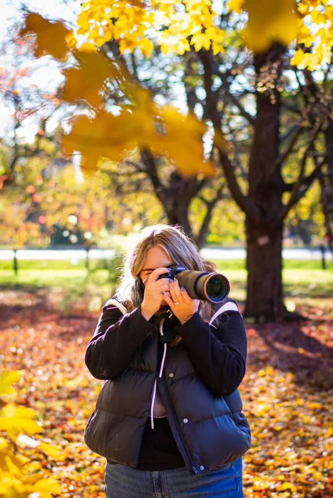 Young woman photographing autumn leaves under golden sunlight in a colorful fall forest.