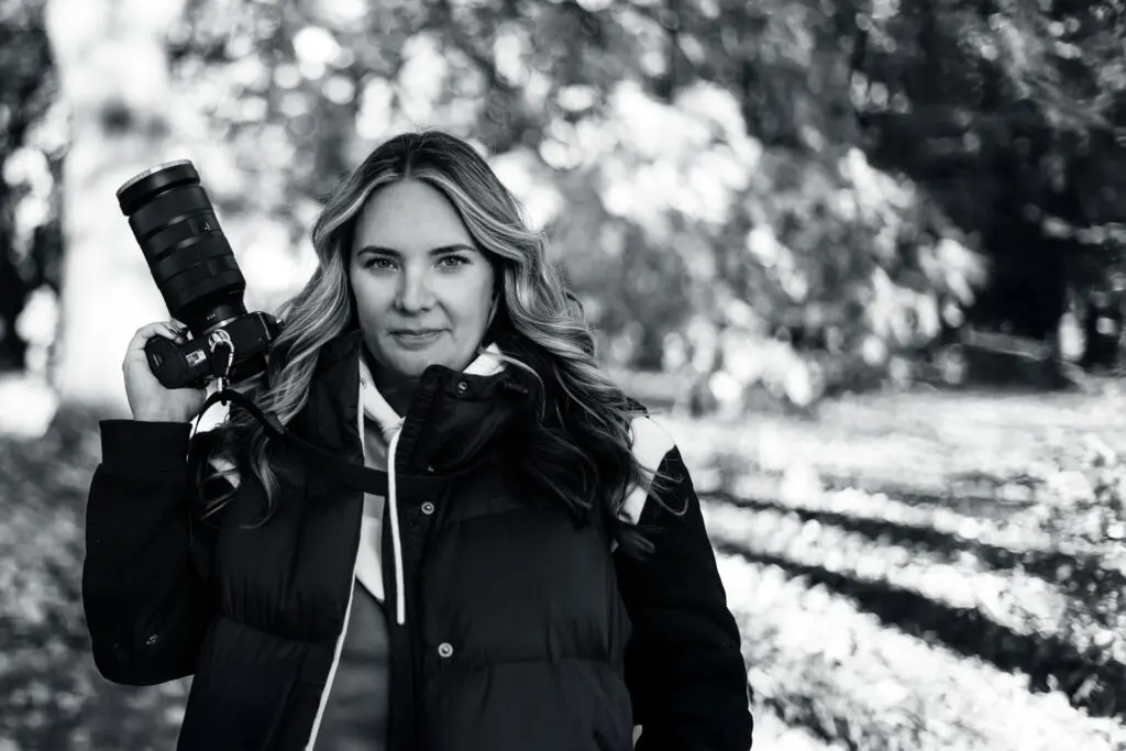 Alicia Riddle confidently holding her camera in a serene black-and-white outdoor forest setting.