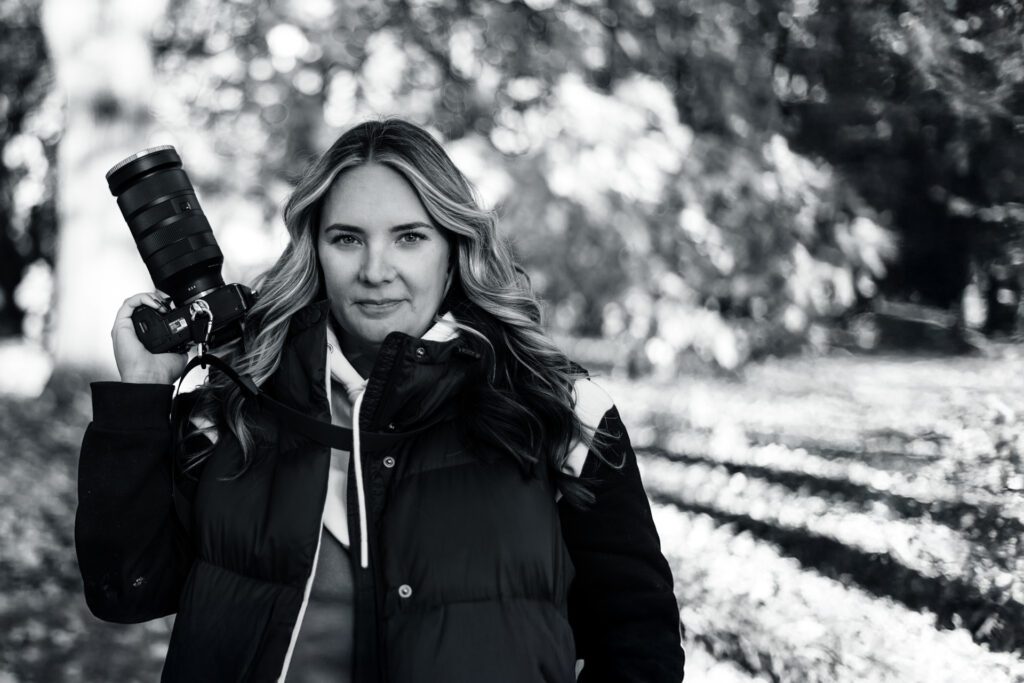 Alicia Riddle confidently holding her camera in a serene black-and-white outdoor forest setting.