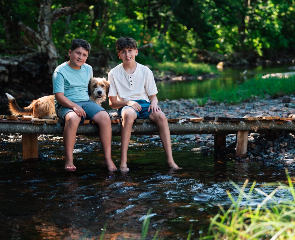 Two barefoot boys and their dog on a wooden bridge over a peaceful forest stream.
