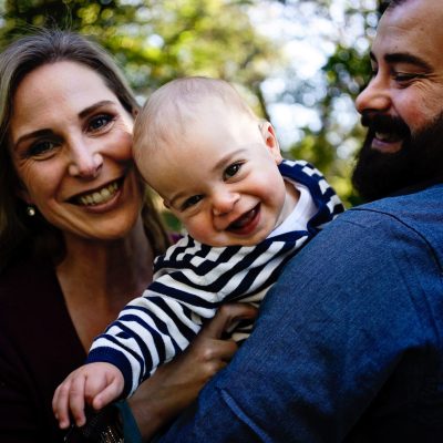 Smiling parents holding their happy baby outdoors under warm sunlight and surrounded by green trees.