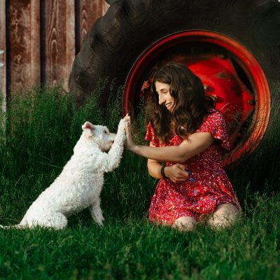 Smiling girl in red floral dress playing with white dog on green countryside lawn.