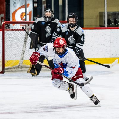 Youth hockey player in white jersey charges puck as opponents defend goal on icy rink.